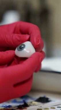 Oftalmolog plastik gözü elinde tutuyor. Detaylı göz modeli. Kırmızı lateks eldivenli ellere odaklan. Dikey video