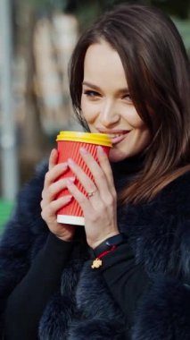 Plastik bardaklı uzun saçlı kız. Kürk mantolu çekici genç kadın şehir parkında kahve içmekten hoşlanıyor. Ağır çekim. Dikey video