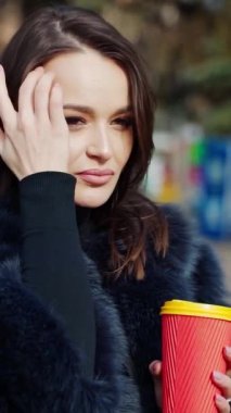 Kışlık ceketli güzel kız. Güzel genç bir kadın parkta plastik bir fincan kahve tutuyor ve gülümsüyor. Ağır çekim. Dikey video