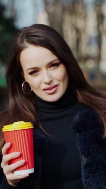Baştan çıkarıcı kadın portresi. Kabarık ceketli güzel bir kız plastik bardak sıcak içecek tutuyor ve şehir parkında kameraya bakıyor. Dikey video