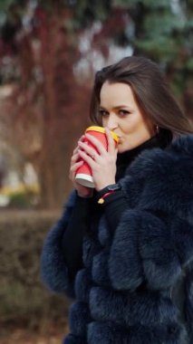 Güzel kız dışarıda kahve içer. Tüylü, sıcak içecekle kameraya bakan şık bir kadının portresi. Parkta şık bir model. Dikey video