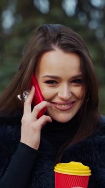 Kabarık ceketli hoş bir kız. Güzel, genç bir esmer cep telefonuyla konuşuyor ve arka planda gülümsüyor. Kürklü kadın dışarıda plastik kahve tutuyor. Dikey video