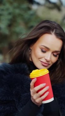 Gülümseyen bir kadının portresi. Kürk mantolu çekici esmer kız parkta plastik bir fincan kahveyle kameraya bakıyor. Ağır çekim. Dikey video