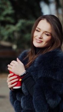 Parkta genç bir kadın var. Zarif kürklü çekici kız elinde plastik bir fincan kahve tutuyor ve dışarıda poz veriyor. Ağır çekim. Dikey video
