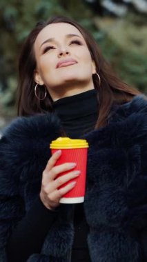 Plastik kahve fincanıyla göz alıcı bir kızın portresi. Parkta dikilip kameraya gülümseyen lüks kürklü çekici genç manken. Ağır çekim. Dikey video
