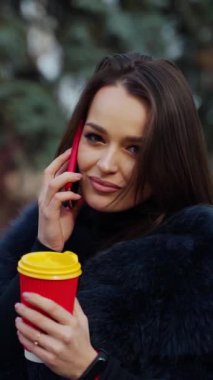 Kürk mantolu çekici bir genç kız. Paltolu güzel bir kadın telefonda konuşuyor ve parkta kahve bardağı tutuyor. Dikey video