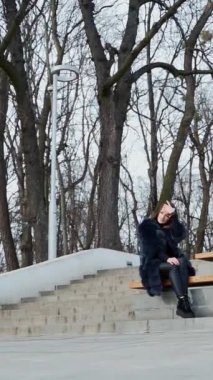 Kız parkta bankta oturuyor. Yapraksız ağaçların arka planında pofuduk ceketli güzel bir kadın tek başına oturuyor. Aşağıdan bak. Dikey video