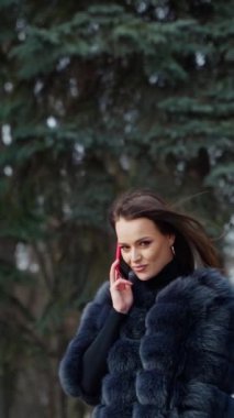 Parkta telefonu olan lüks bir kız. Kürklü çekici bir kadının portresi parkta yürüyor ve cep telefonuyla konuşuyor. Ağır çekim. Dikey video