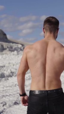 Beyaz Kanyon 'da güneşli bir günde atlet. Sporcunun doğadaki arka görünüşü. Gömleksiz vücut geliştirici dağlarda siyah arabaya doğru yürüyor. Ağır çekim. Dikey video