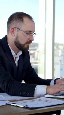 Dizüstü bilgisayar kullanan ciddi bir erkek işçi. Büyük pencereli bir ofiste dizüstü bilgisayarla çalışan odaklanmış bir iş adamı. Bilgisayarda daktilo eden gözlüklü bir iş adamı. Dikey video