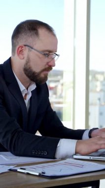 Dizüstü bilgisayar kullanan ciddi bir erkek işçi. Büyük pencereli bir ofiste dizüstü bilgisayarla çalışan odaklanmış bir iş adamı. Bilgisayarda daktilo eden gözlüklü bir iş adamı. Dikey video