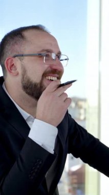 Ofisten biriyle konuşan gülümseyen bir iş adamı. Gözlüklü ve kostümlü girişimci portresi şehrin penceresinin arkasında masasında oturuyor. Dikey video