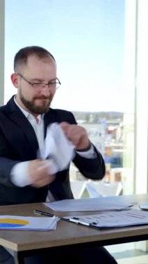 Sinirli iş adamı ofiste. Sinirli girişimci adam masada oturuyor ve kağıtları pencerenin arkasına fırlatıp yırtıyor. Dikey video