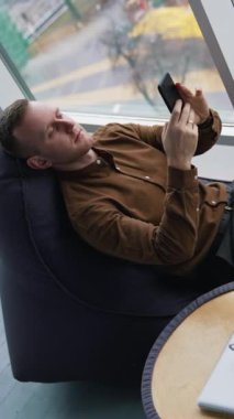 Adam telefonu olan yumuşak bir sandalyede dinleniyor. Şehir ulaşımı geçmişi olan ve cep telefonunu kullanan genç bir adam pencerenin yanında oturuyor. Küçük masada bir fincan kahve. Üst Manzara. Dikey video