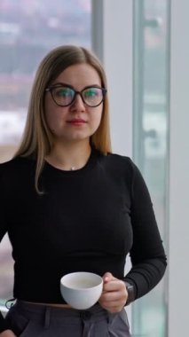 Kapalı mekanda bir fincan kahvesi olan iş kadını. Gözlüklü genç bir kadın ofisin penceresinin yanında durup sıcak içecek içerken mola veriyor. Dikey video