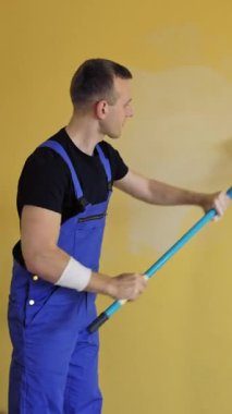 Adam evdeki duvarları boyuyor. Mavi üniformalı genç inşaatçı uzun saplı boya silindirli duvarları yeniliyor. Resim hizmetleri. Dikey video