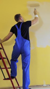 İnşaat merdiveninin yakınındaki duvarları boyayan bir zanaatkar. Profesyonel boyacı mavi tulumlu. Odada boya silindiri ile tadilat yapıyor. Dikiz aynası. Dikey video