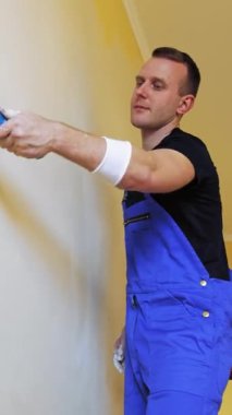 Boyacı duvarları silindirle boyuyor. Tulumlu genç bir işçinin portresi. Odada yeni bir renkle makyaj yapıyor. Profesyonel resim servisi. Dikey video