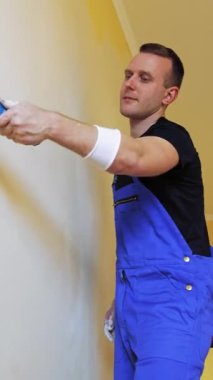 Boyacı duvarları silindirle boyuyor. Tulumlu genç bir işçinin portresi. Odada yeni bir renkle makyaj yapıyor. Profesyonel resim servisi. Dikey video