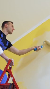 Duvarları içeride boyayan profesyonel bir zanaatkar. Mavi tulumlu genç erkek işçi bir bina merdiveninin üzerinde duruyor ve boya silindiri kullanıyor. Aşağıdan bak. Dikey video