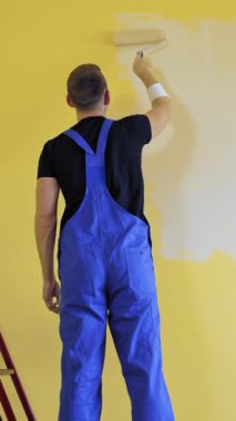 Kapalı alanda makyaj yapan bir ressam. Merdivenin yanında boya silindiri olan duvarı boyayan bir erkek işçi. Odanın iç kısımlarında gelişme var. Arkadan bak. Dikey video