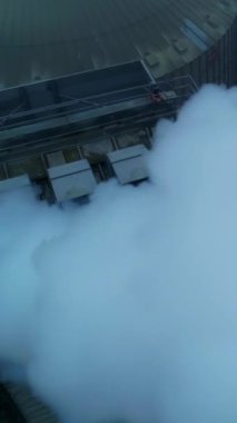Asansör fabrikasında çalışan, kalın borular, yoğun duman çıkaran. Gri havanın arka planında ambar ambarının tepesinde uçuyor. Dikey video