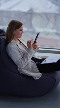 Sakin sakin telefonuna bakıyor sandalyede oturuyor ve önünde bacaklarını açıyor. Kadının dizüstü bilgisayarı ve kucağında gözlükleri var. Arka planda kar pencerenin arkasına düşüyor. Dikey video