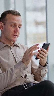 Akıllı telefonda görüntülü konuşma. Orta yaşlı bir adam konuşur, gülümser ve elini pencerenin yanındaki sandalyede sallar. Arkaplan bulanık. Dikey video