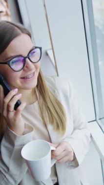 Gülümseyen genç bayan elinde bir bardakla panoramik pencerenin önünde duruyor. Kadın telefonda konuşuyor ve çay içiyor. Yüksek açı. Dikey video