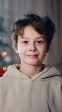 Kameraya gülümseyen bej kapüşonlu yakışıklı çocuk. Koyu saçlı genç kapalı bir Noel ağacının bulanık zemininde poz veriyor..