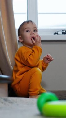 Pencerenin yanında oturan küçük bir çocuk ve içerideki merdivenler gözlerini ovuşturuyor. Bebek parmağını ağzına çeker ve odaya bakar..