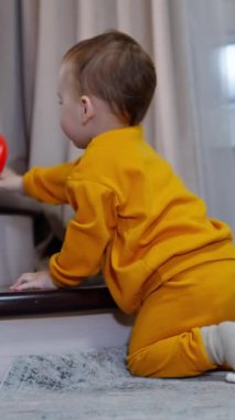 Turuncu spor takım elbiseli sevimli çocuk merdivenlerin yanında yerde oturuyor. Çocuk oyuncağı atar ve yuvarlanmasını izler..