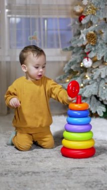 Turuncu spor takım elbiseli sevimli küçük çocuk, tatil için dekore edilmiş odada oyuncak piramit inşa ediyor. Arkaplanda Noel ağacı.