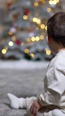 Sevimli bebek sırtüstü yere oturmuş Noel ağacına bakıyor. Çocuk hızla ağaca tırmanıyor..