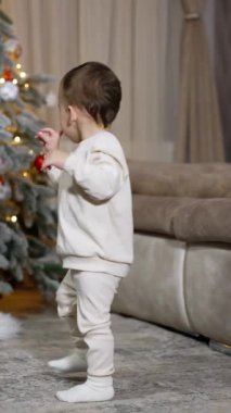 Beyaz elbiseli tatlı çocuk odada kameraya doğru duruyor. Bebek birkaç oyuncak almak için Noel ağacına yaklaşıyor..