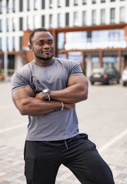 Kaslı bir adam günlük spor kıyafetleriyle dışarıda rahatça poz veriyor. Kolları çapraz duran kaslı bir birey modern bir açık hava alanında kendine güvenini gösteriyor..