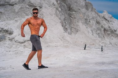 Açık mavi gökyüzünün altındaki kumlu arazide güvenle poz veren bir adam. Formda bir adam, beyaz kum ve mavi gökyüzünün arka planında poz verir..