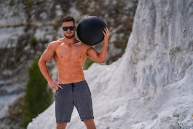 Gündüz vakti kayalık arazide tıbbi topla egzersiz yapan bir adam. Tişörtsüz bir adam, kayalık oluşumların yanında bir tıp topuyla güvenle poz verir..