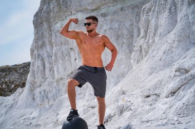 Genç adam yazın kayalık bir sahilde poz verirken kaslarını esnetir. Üstsüz genç bir adam gururla kaslarını taşlı bir zemine karşı gösterir..