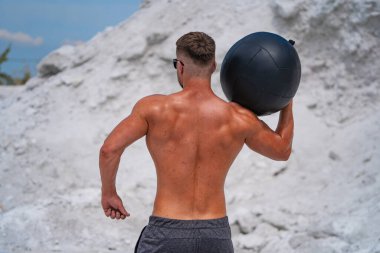Adam kum tepeciğinin yanında ağırlık topuyla dışarıda egzersiz yapıyor. Tişörtsüz bir adam dışarıda antrenman yaparken ağır bir topu kaldırır..