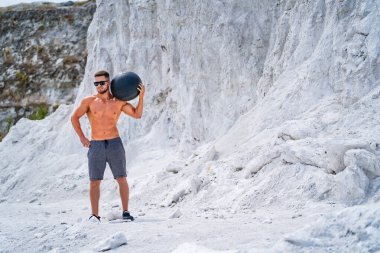 Siyah bir ilaç topuyla dışarıda tebeşir gibi bir yerde egzersiz yapan bir adam. Bir adam dışarıda antrenman yapıyor, tebeşir gibi bir arazide dururken elinde bir ilaç topu tutuyor..