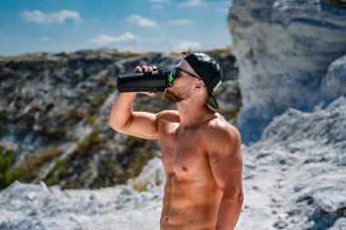 Kayalık bir arazide siyah bir şişeden su içen adam. Üstsüz bir adam, siyah bir su şişesinden kayalık bir zemine doğru su içmekten zevk alır..