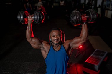 Vücut geliştirici akşam antrenmanı sırasında spor salonunda ağır dambıllar kaldırıyor. Vücut geliştiren biri, loş bir spor salonunda dambıllarla üstten baskı yapıyor..