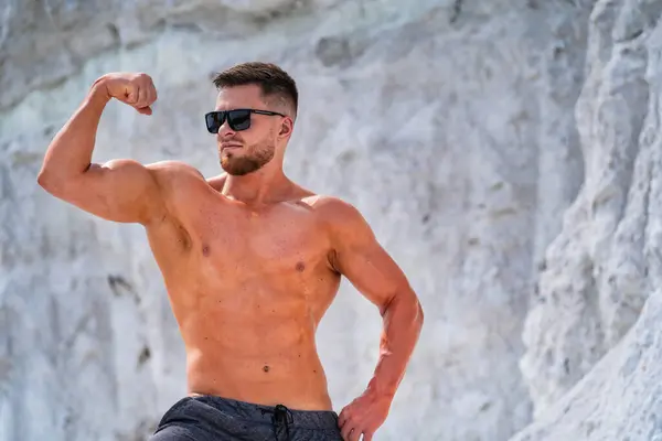 stock image Young man flexes muscles on a rocky beach during sunny weather. A shirtless man shows off his physique against a rugged, light-colored backdrop.