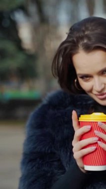Kürklü, açık havada içkisi olan güzel bir kız. Bulanık park arka planında elinde plastik bardakla güzel bir kadının portresi. Dikey video