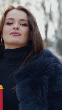 Çekici kadın elinde plastik bardakta kahve tutuyor. Kürk mantolu güzel bir kızın portresi şehir parkında eli olan birini arıyor. Ağır çekim. Dikey video