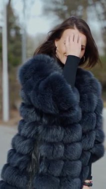 Modaya uygun bir kadının sokak portresi. Parkta yürüyen şık siyah kürk giyen genç ve güzel bir manken. Kameraya poz ver. Sokak modası konsepti. Dikey video