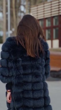 Parkta poz veren çekici bir kız. Siyah kürklü güzel bir kadın kameraya bakıyor ve dışarıda gülümsüyor. Ağır çekim. Dikey video