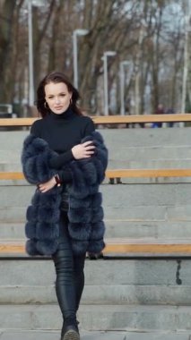 Sonbahar parkında güzel bir model poz veriyor. Siyah kürklü genç bir kadın yürüyor ve kameraya bakıyor. Ağır çekim. Dikey video