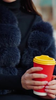 Kadın elleri dışarıda bir bardak içeceği tutuyor. Kürk mantolu güzel bir kadın parkta oturuyor ve plastik camdan içiyor. Ağır çekim. Dikey video